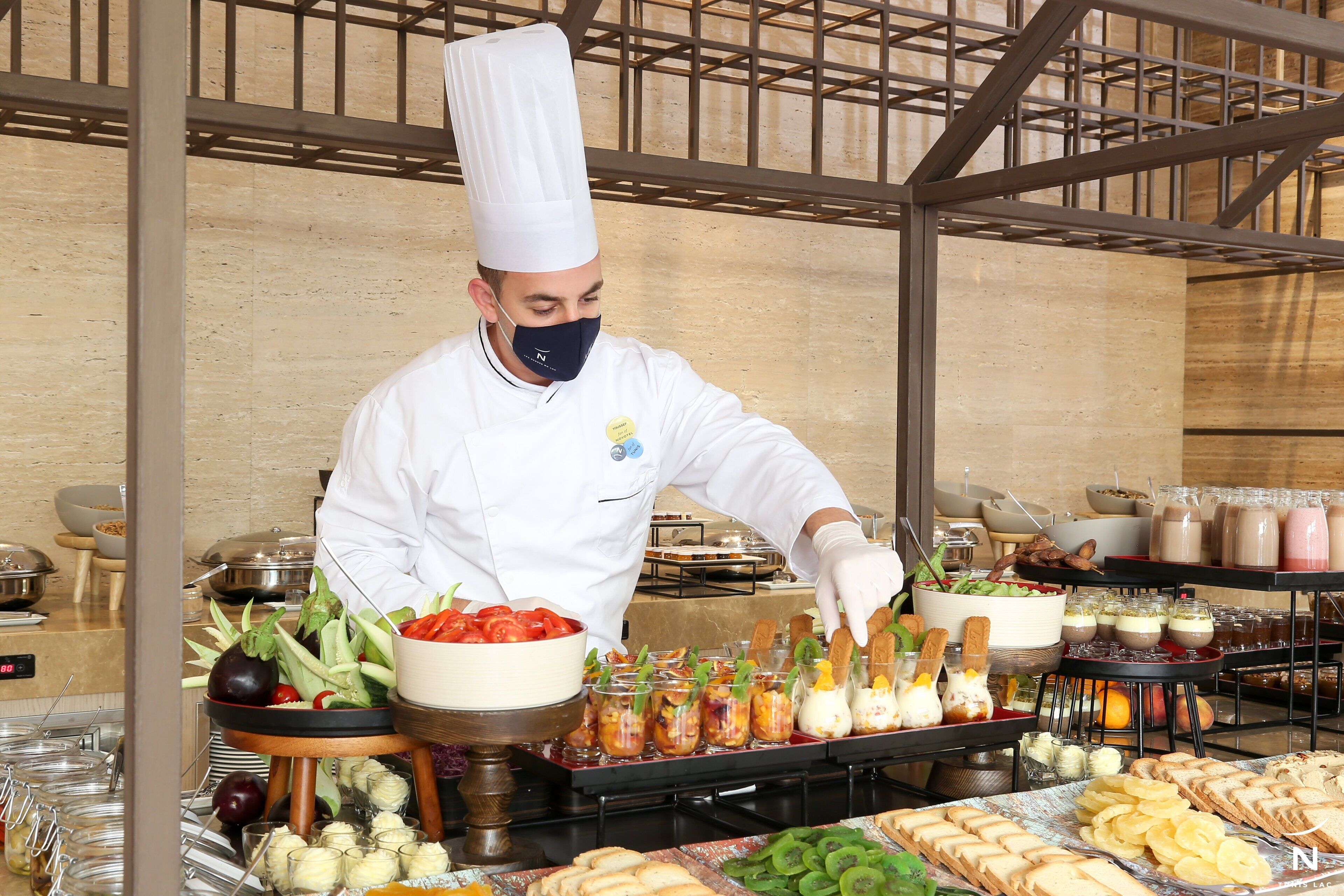 A chef in a white uniform and hat preparing food in a kitchen with various dishes and ingredients visible.