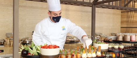 A chef in a white uniform and hat preparing food in a kitchen with various dishes and ingredients visible.