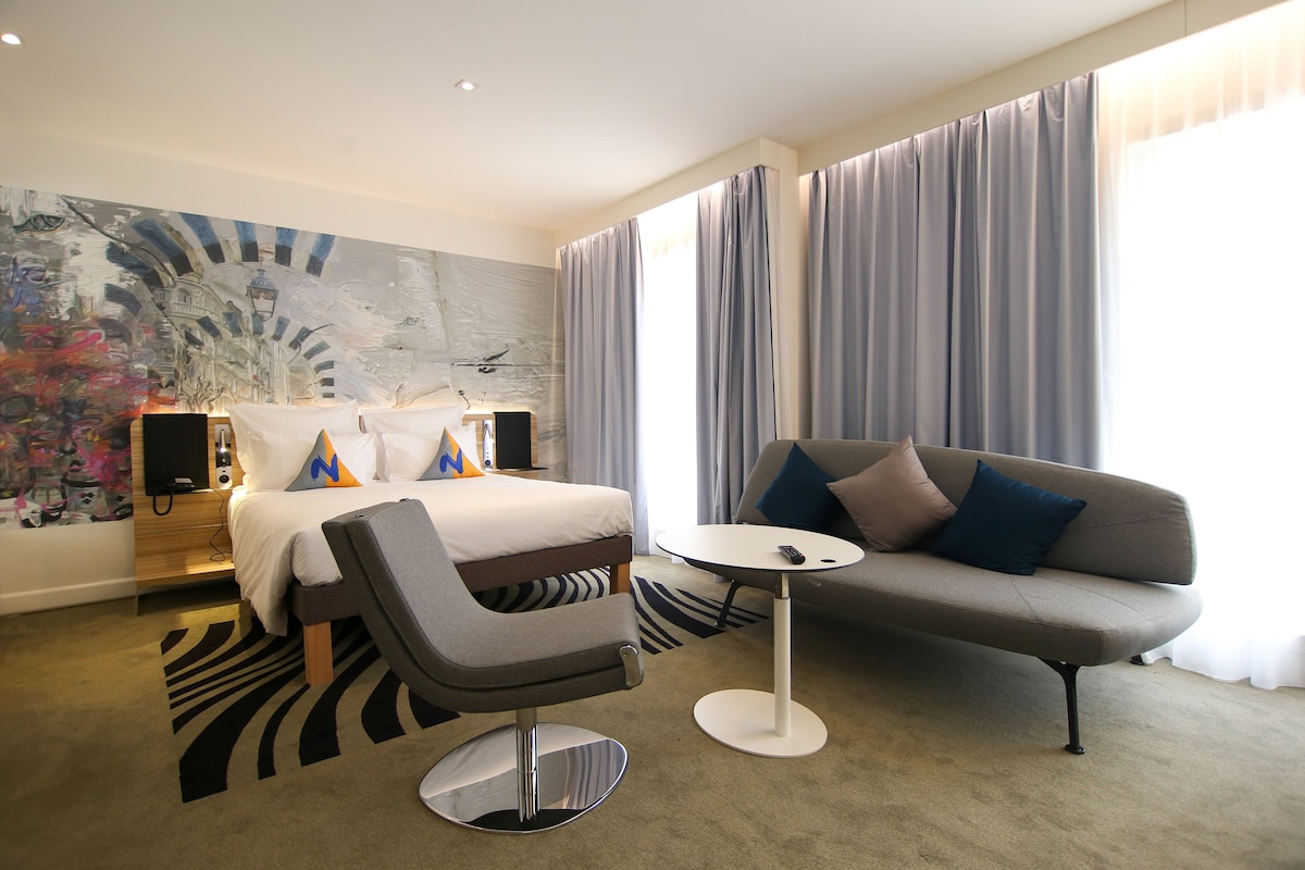 A modern hotel room with a bed, a grey sofa, a small round table, and a large abstract painting on the wall.