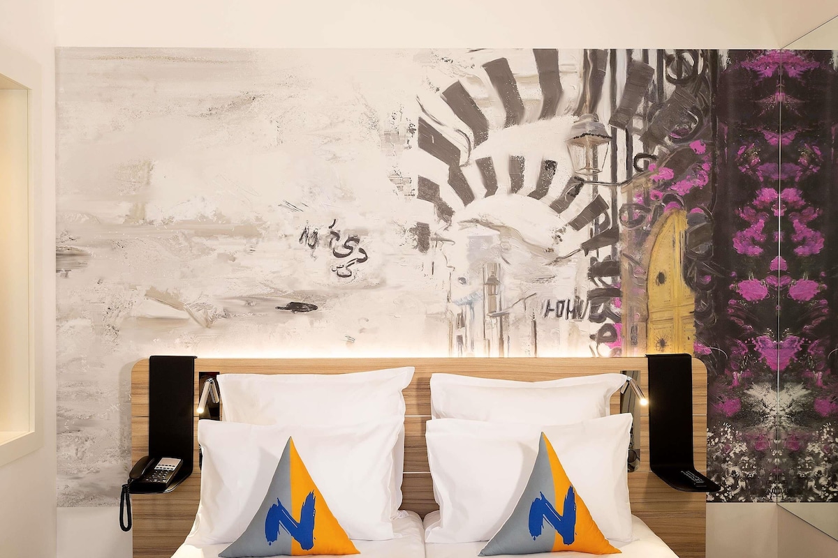 A hotel room with a bed, two pillows with a blue and orange design, a phone on the nightstand, and a wall with abstract art.