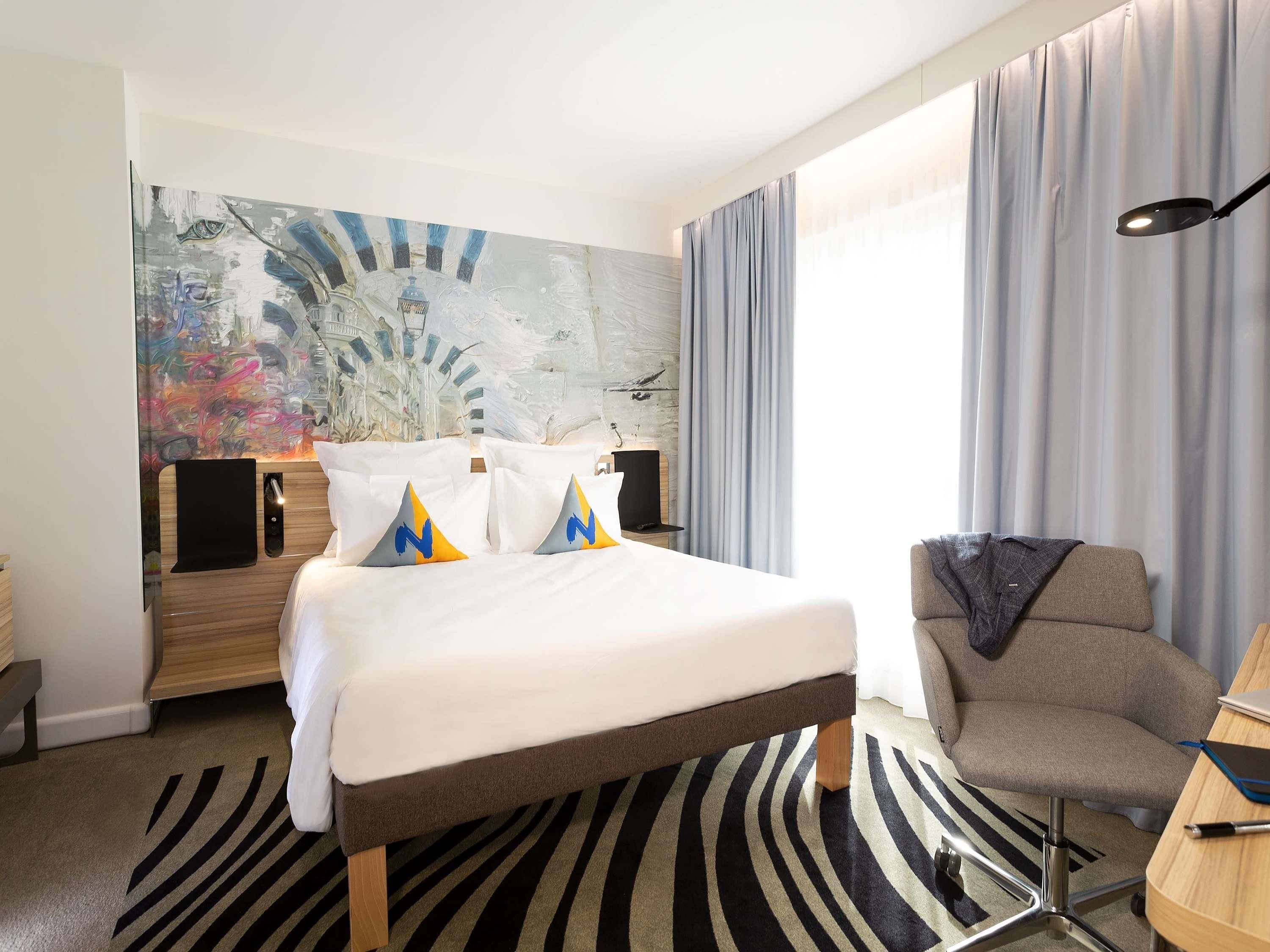 A hotel room with a bed, a desk, a chair, a lamp, a painting on the wall, and a window with curtains.