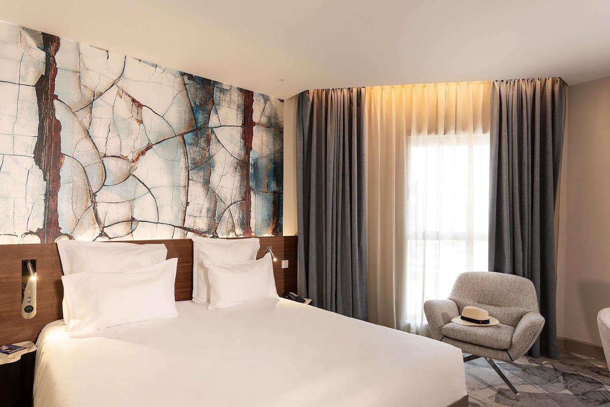A hotel room with a large bed, a nightstand, a chair, a wall art piece, and a window with curtains.