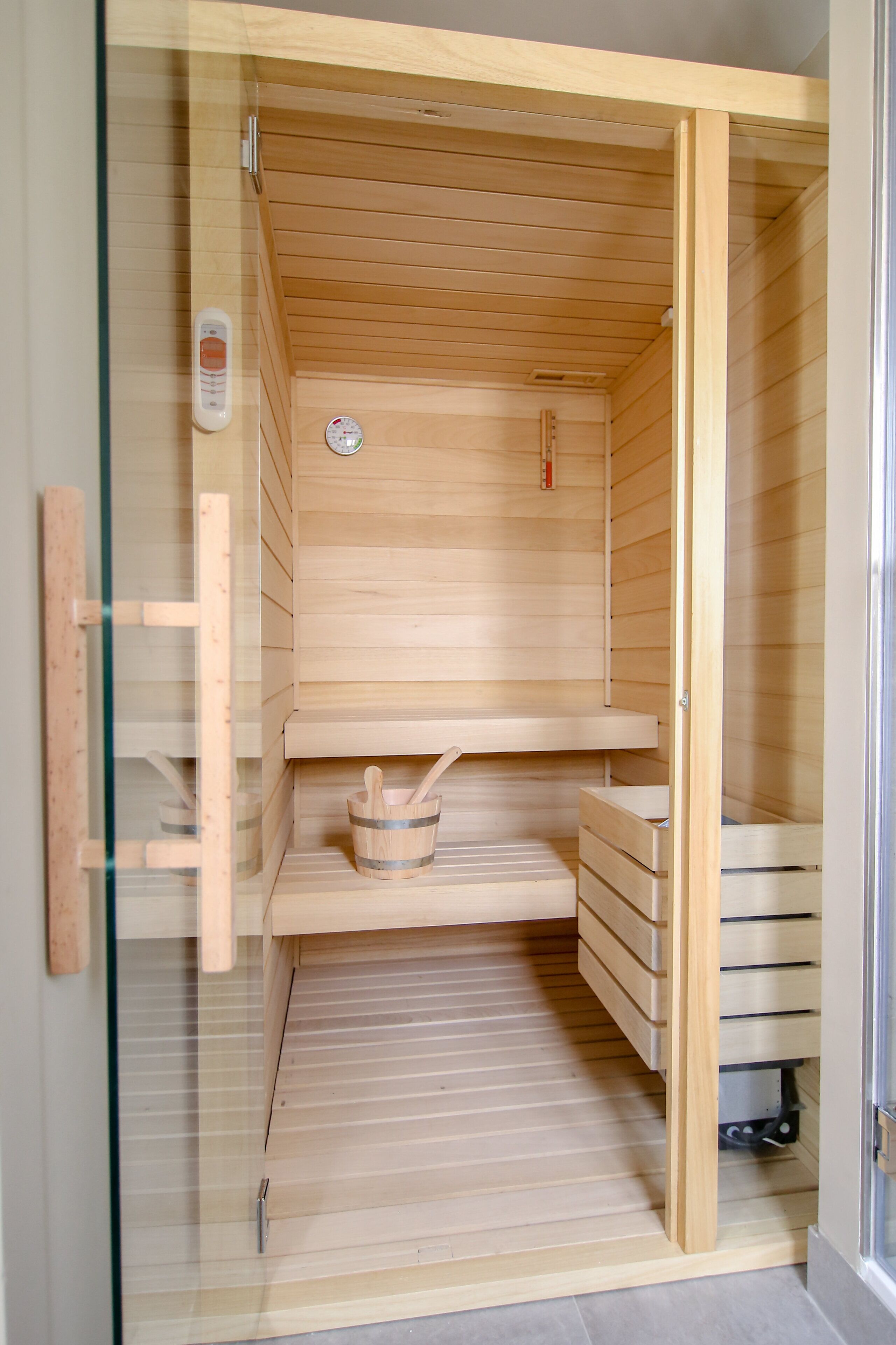 A sauna with wooden walls and a glass door, featuring a control panel, a small shelf, and a wooden bench.