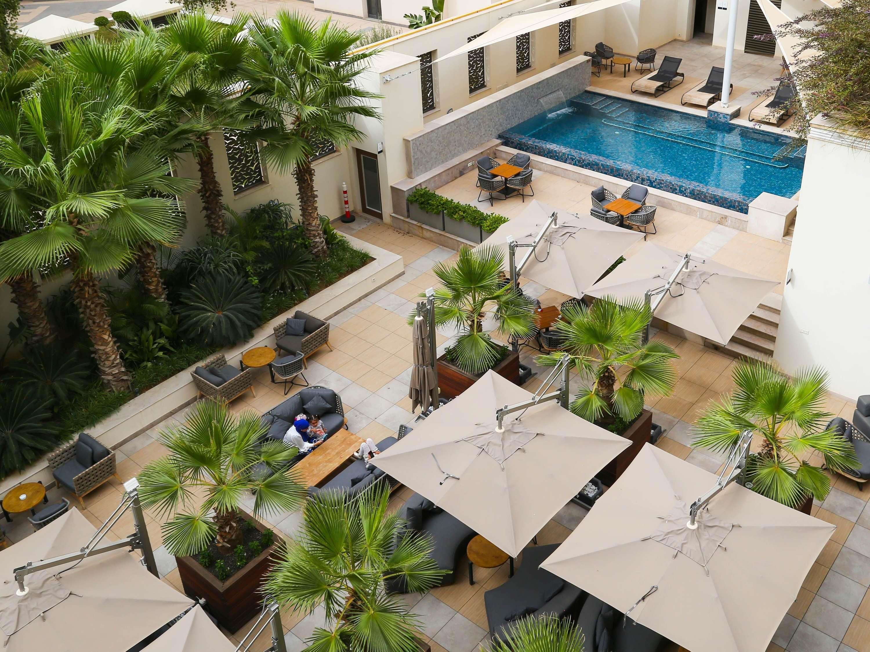 A rooftop pool area with lounge chairs, umbrellas, and palm trees.