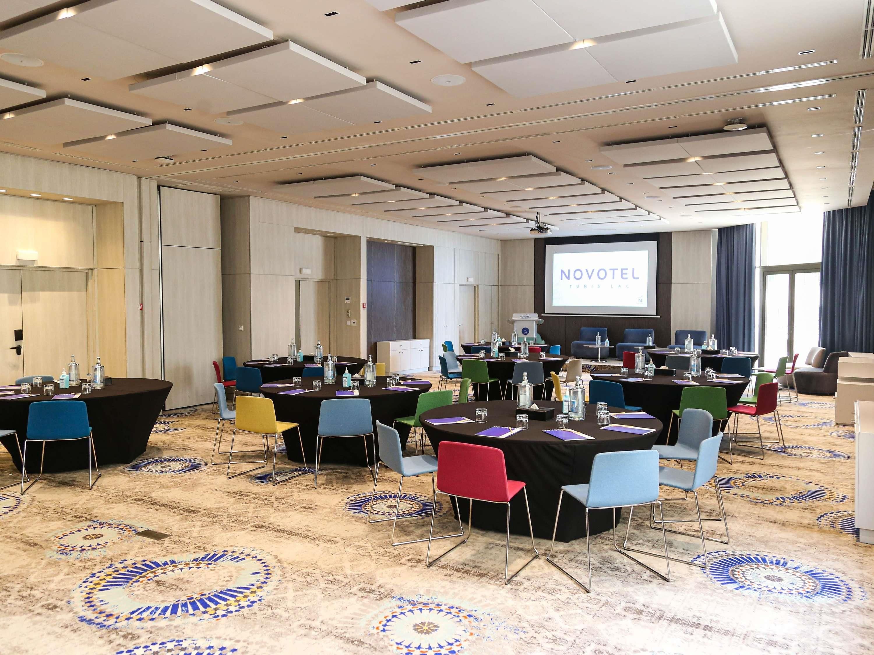 Un salón de conferencias con varias mesas y sillas, una pantalla de proyector mostrando 'NOVOTEL' y un suelo alfombrado con un diseño circular.