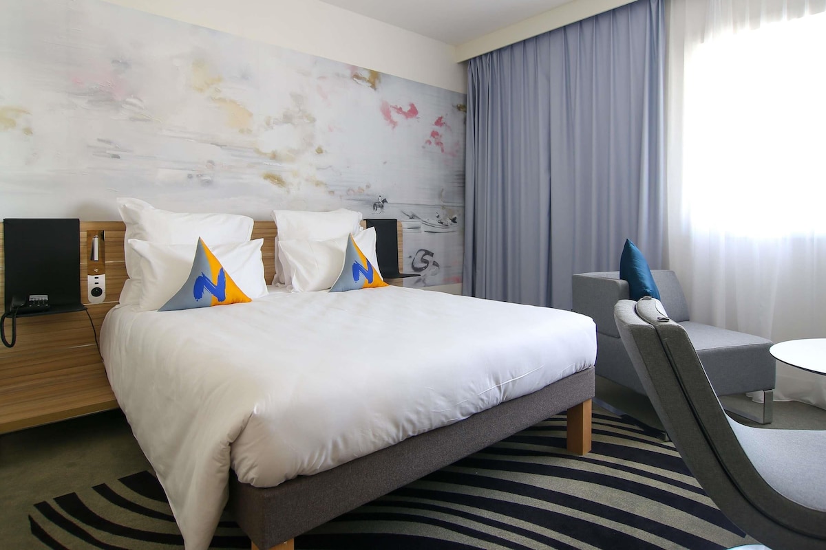 A hotel room with a large bed, a desk with a chair, a flat-screen TV, and a window with curtains.