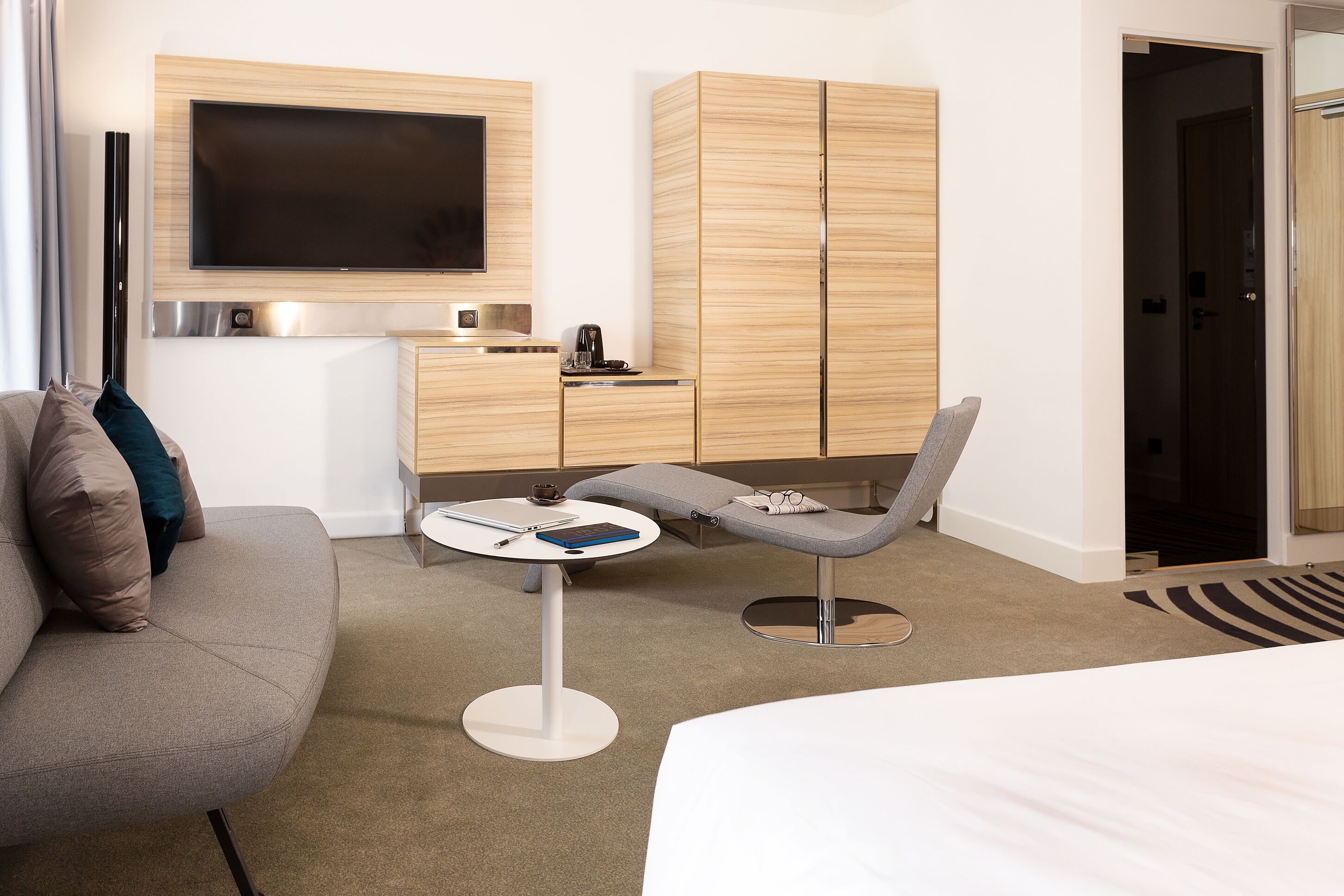 Et moderne hotelværelse med et fladskærms-tv, en træskab, en grå lænestol, et lille rundt bord og en seng.