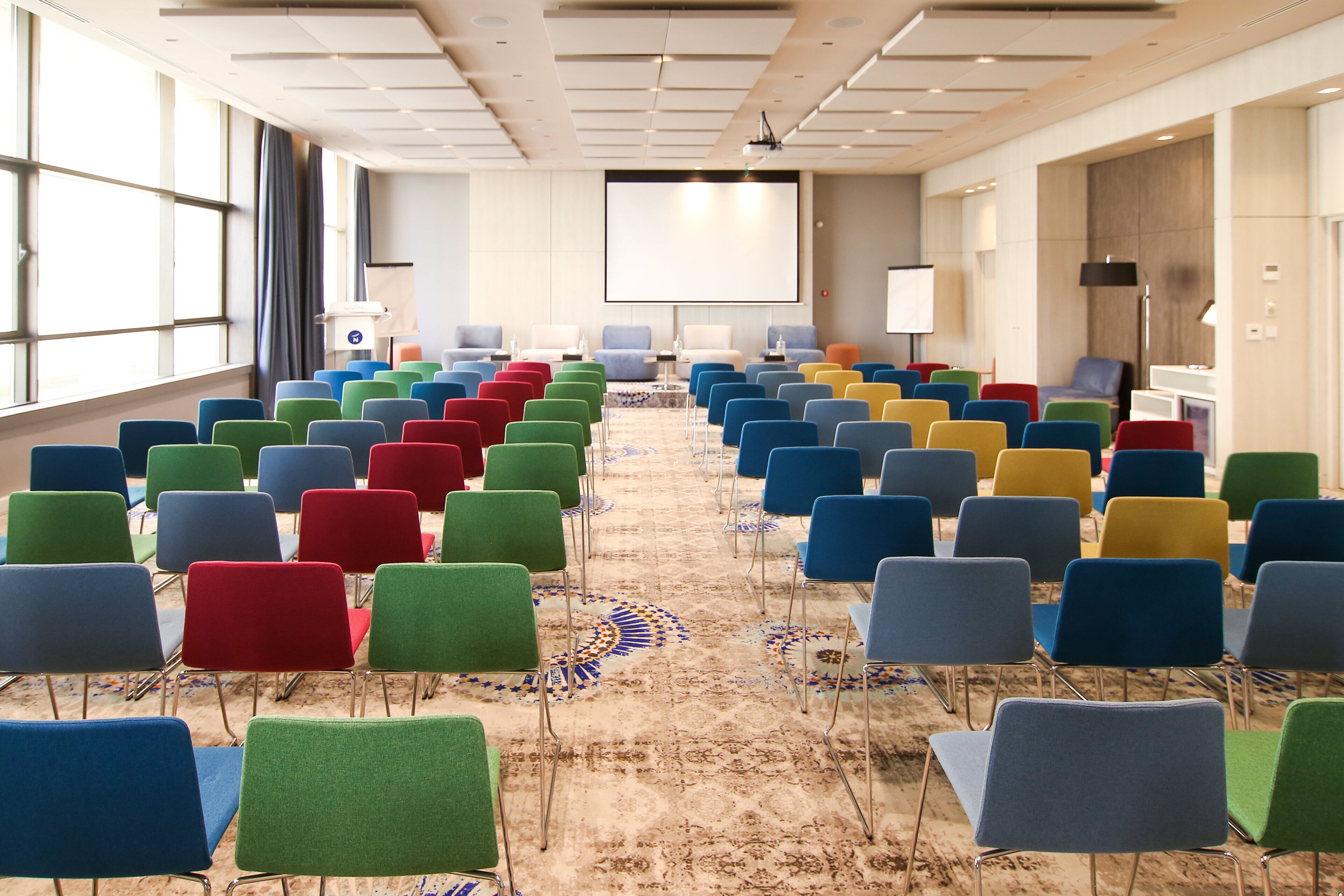 A conference room with rows of colorful chairs, a projector screen, and a carpeted floor.