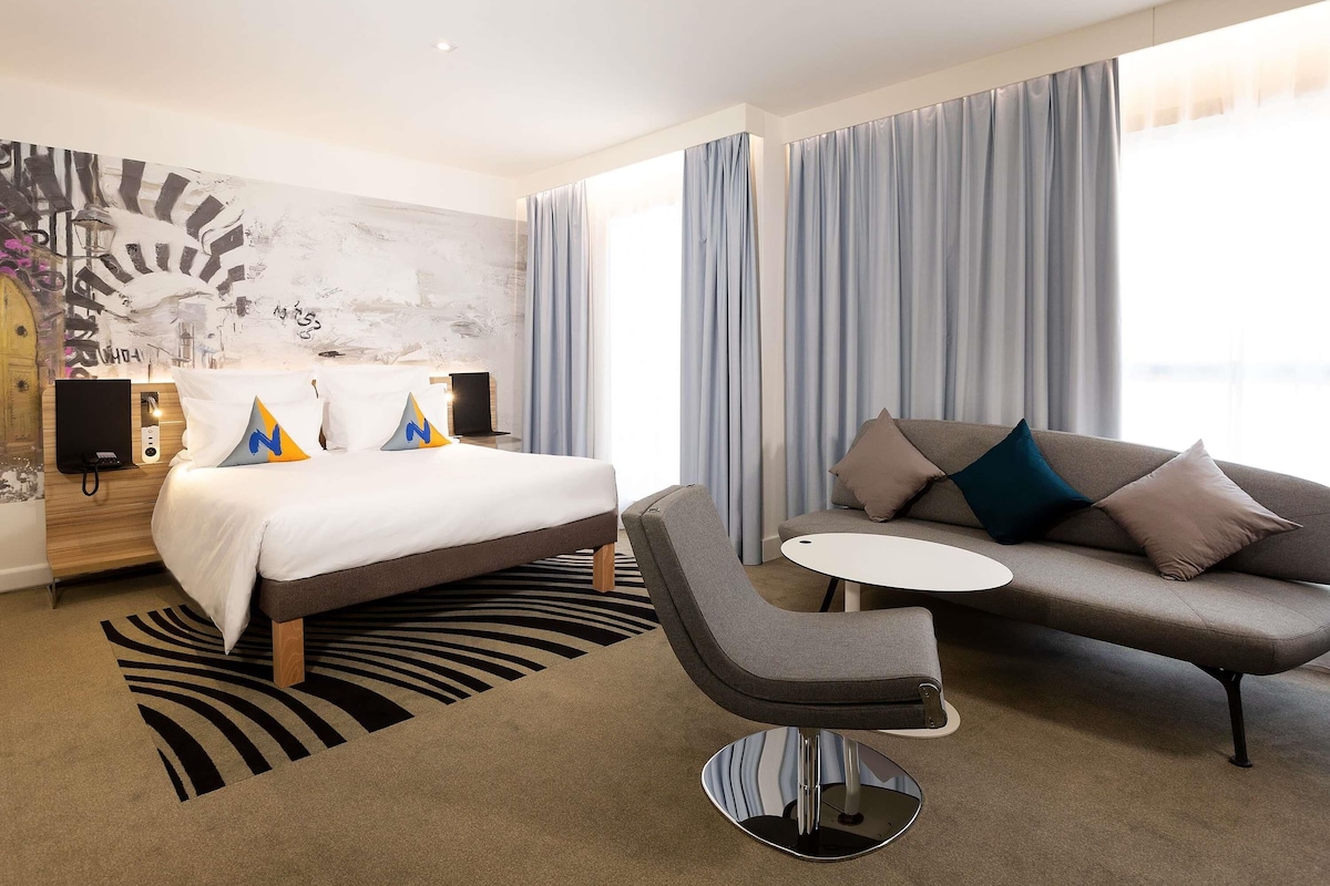 A hotel room with a bed, a sofa, a small table, a chair, a wall with a mural, and a window with curtains.