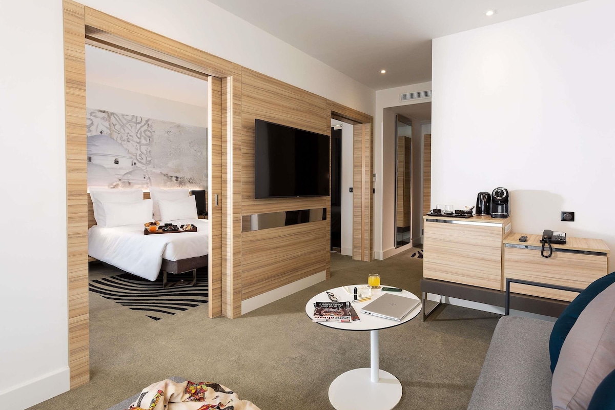 A hotel room with a bed, a TV, a desk with a phone and coffee maker, and a small table with a laptop and magazines.
