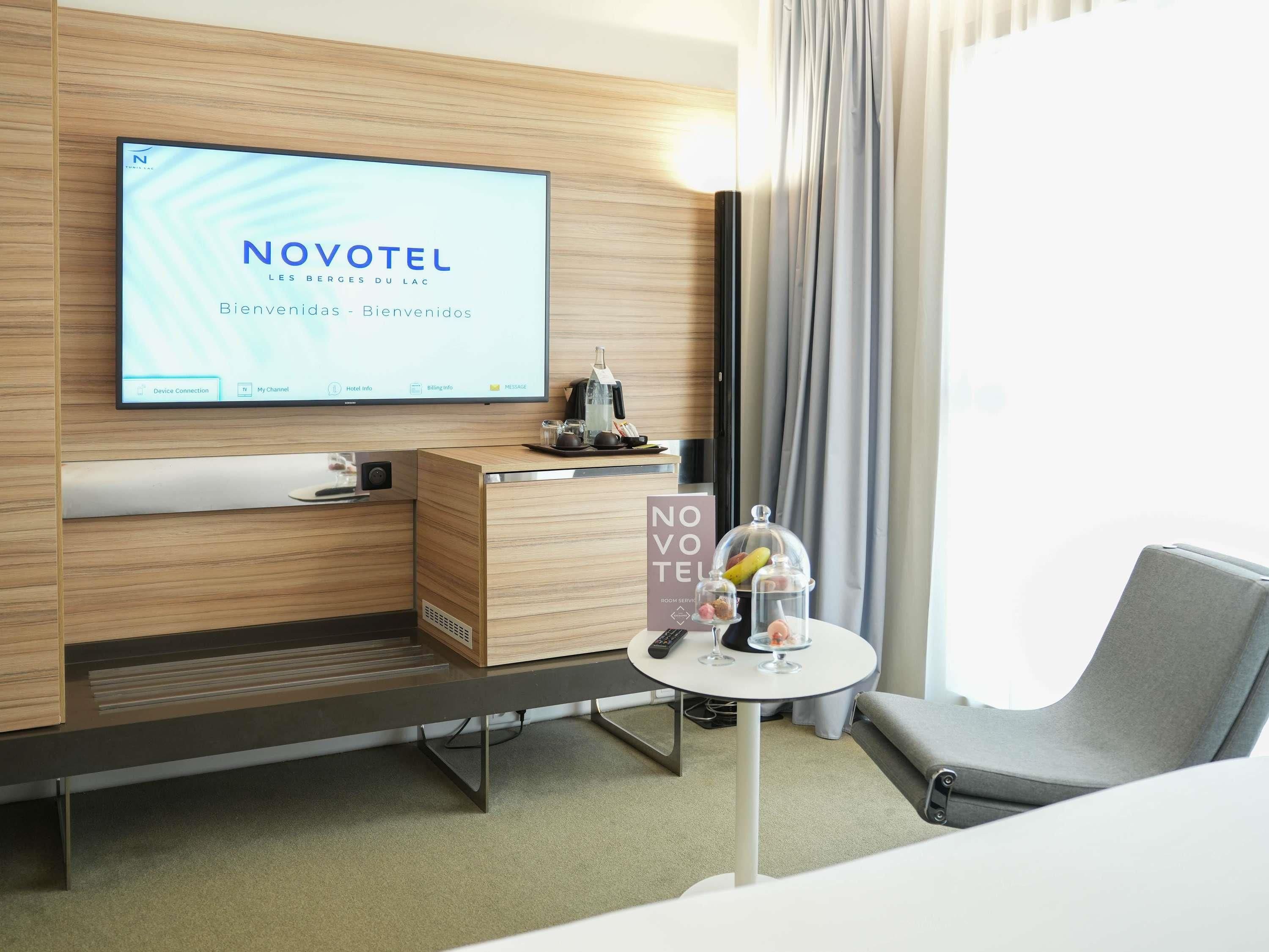 A hotel room with a television displaying 'NOVOTEL' and a sign indicating 'ROOM SERVICE.'