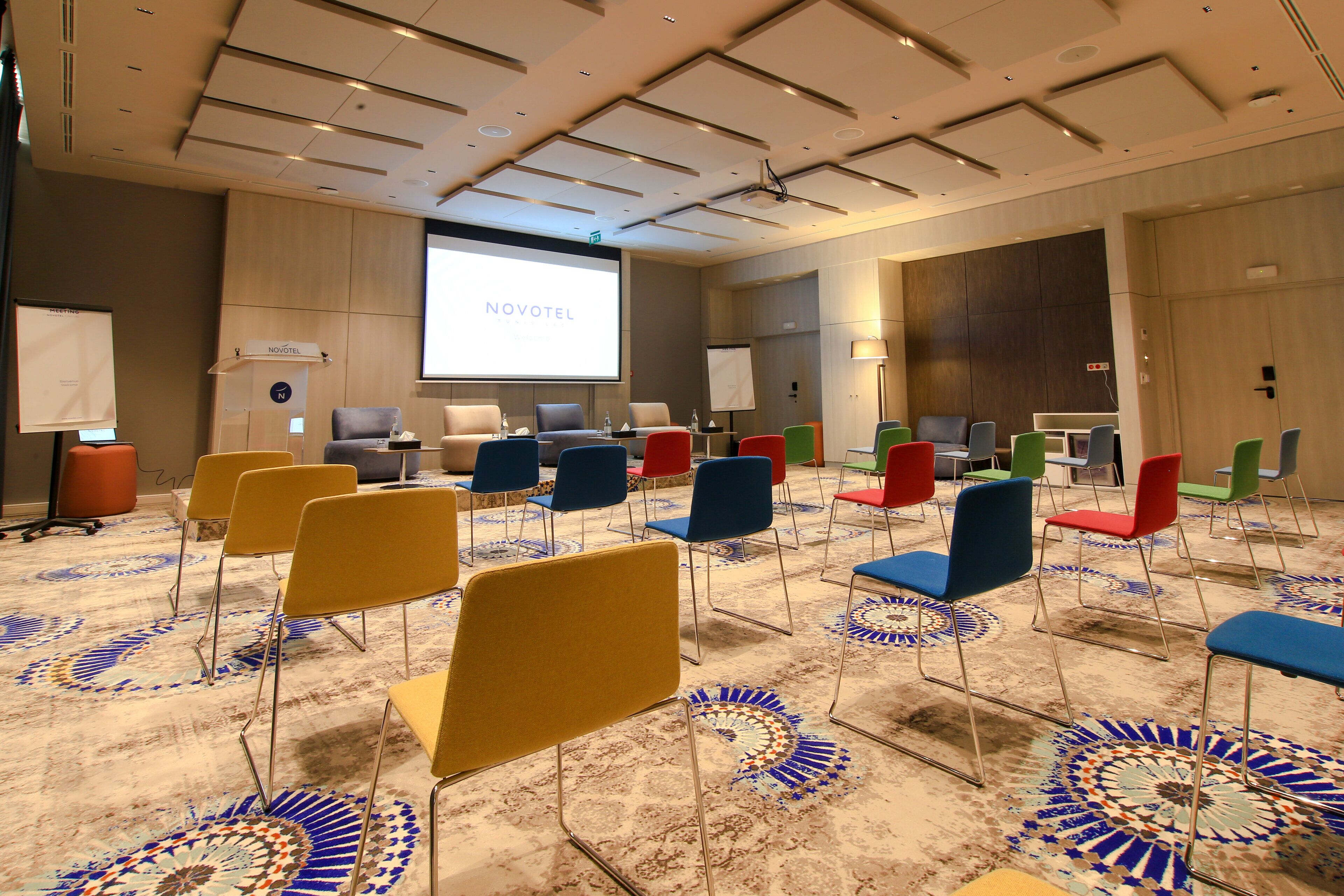 A conference room with a projector screen displaying "NOVOTEL," rows of colorful chairs, and a carpet with a circular pattern.