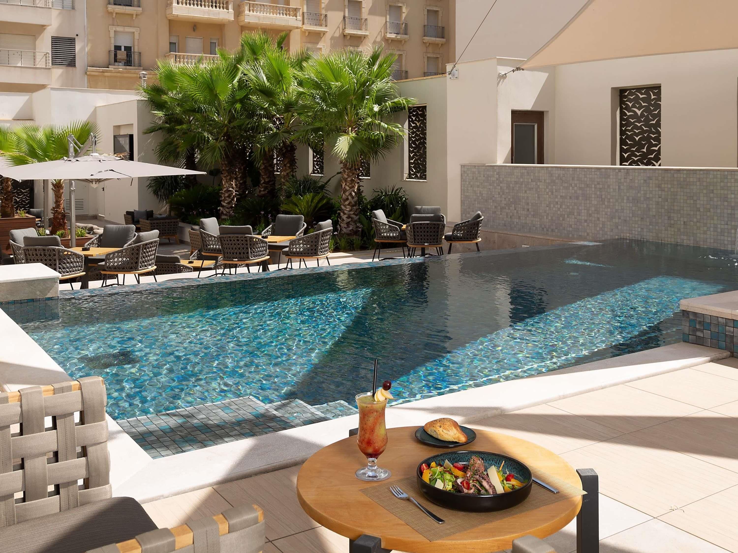 A poolside area with lounge chairs, a dining table with a meal, and a view of a building with balconies.