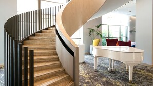 A modern spiral staircase with wooden steps and black railings, leading to a white piano in a well-lit room with large windows and a patterned carpet. - Novotel Tunis Lac (Tunis)