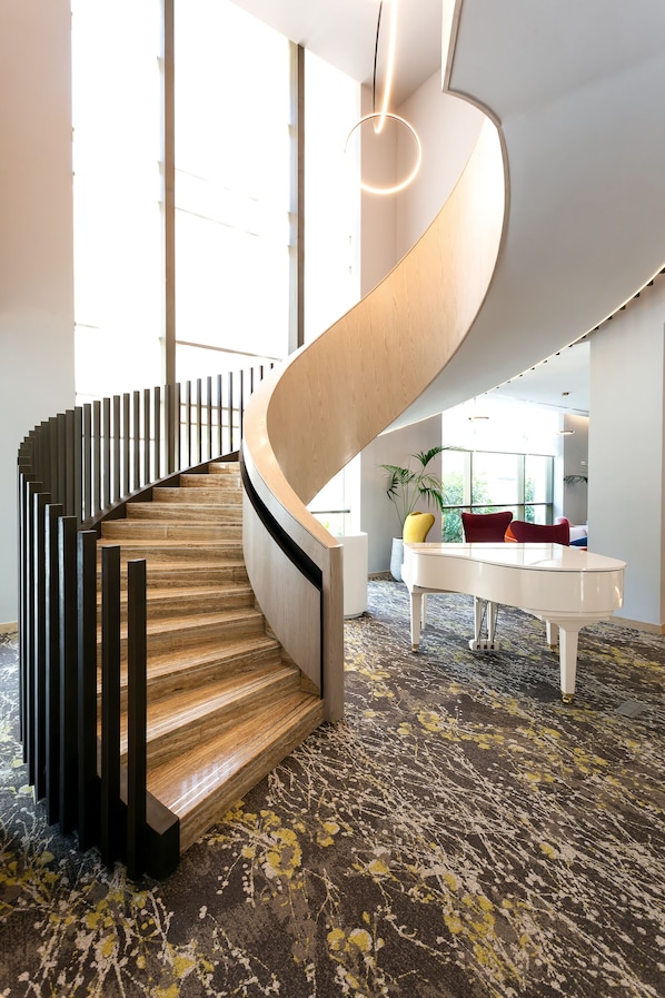 Eine moderne Wendeltreppe mit Holzbodenstufen und schwarzen Geländern, die in einen gut beleuchteten Raum mit großen Fenstern und einem gemusterten Teppich zu einem weißen Klavier führt.