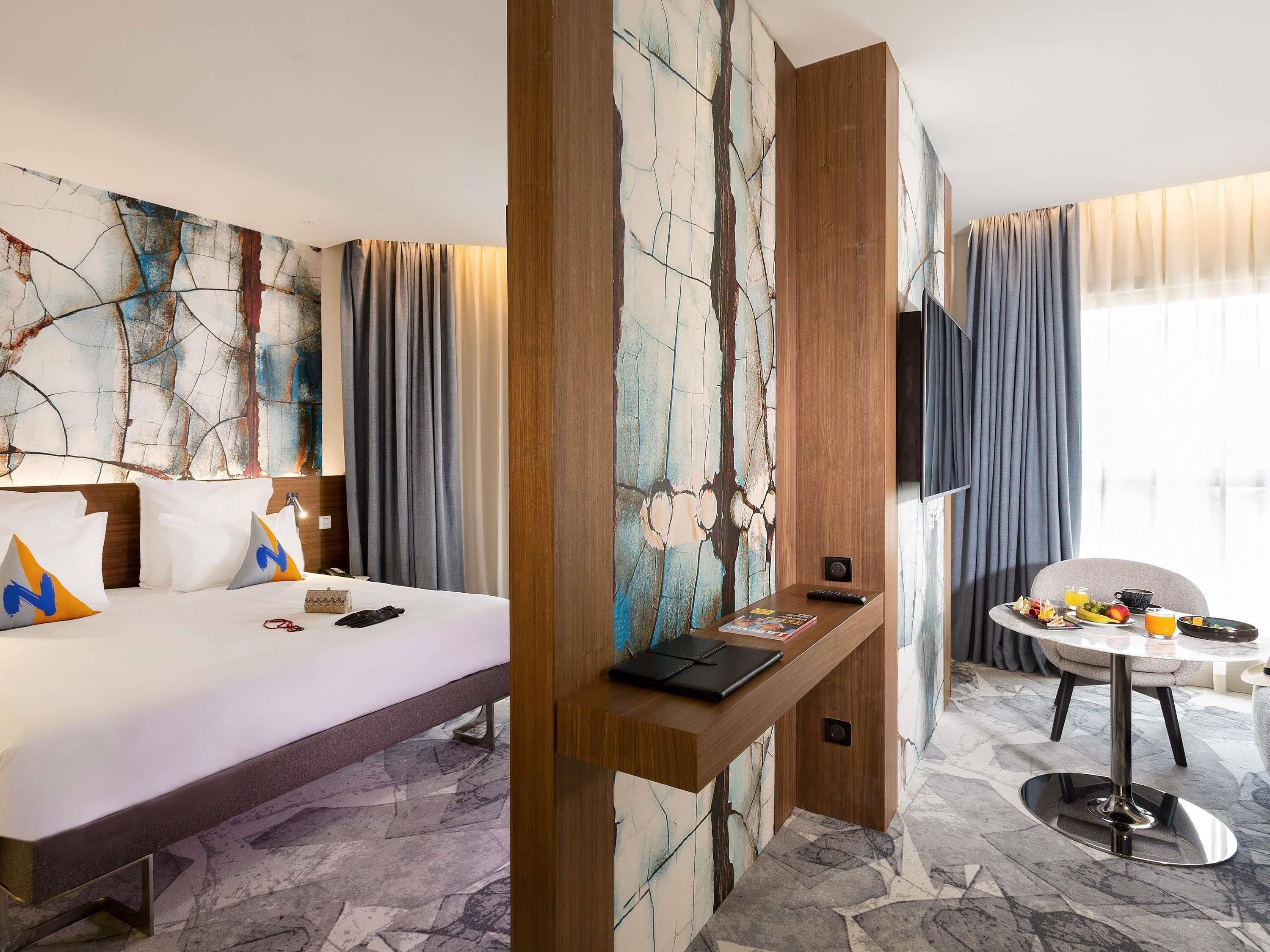 A hotel room with a bed, a desk, a chair, a TV, and a dining table with breakfast items.