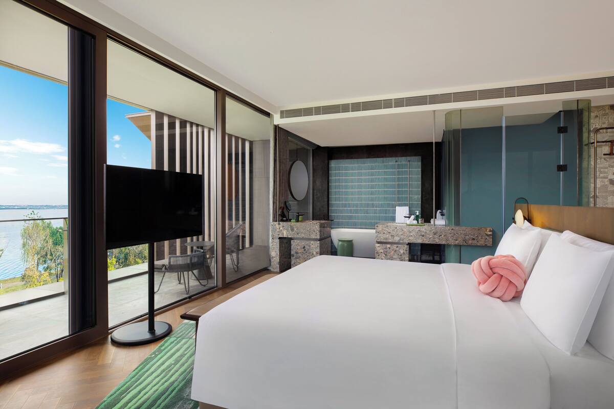 A modern hotel room with a large bed, a TV, a balcony with a view of the water, and a bathroom with a mirror and sink.