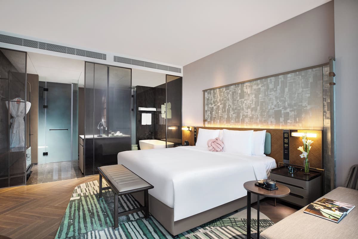 A modern hotel room with a large bed, a desk, a chair, a small table, a nightstand, and a bathroom with a shower and sink.