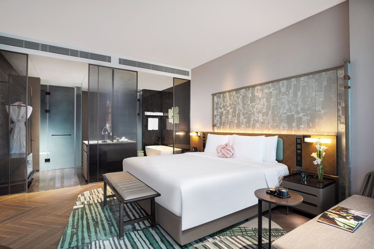 A modern hotel room with a large bed, a desk, a chair, a small table, a nightstand, and a bathroom with a shower and sink.