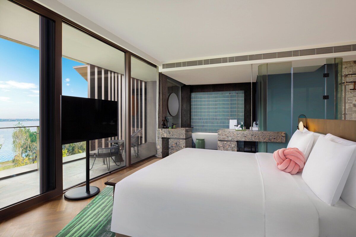 A modern hotel room with a large bed, a TV, a balcony with a view of the water, and a bathroom with a mirror and sink.