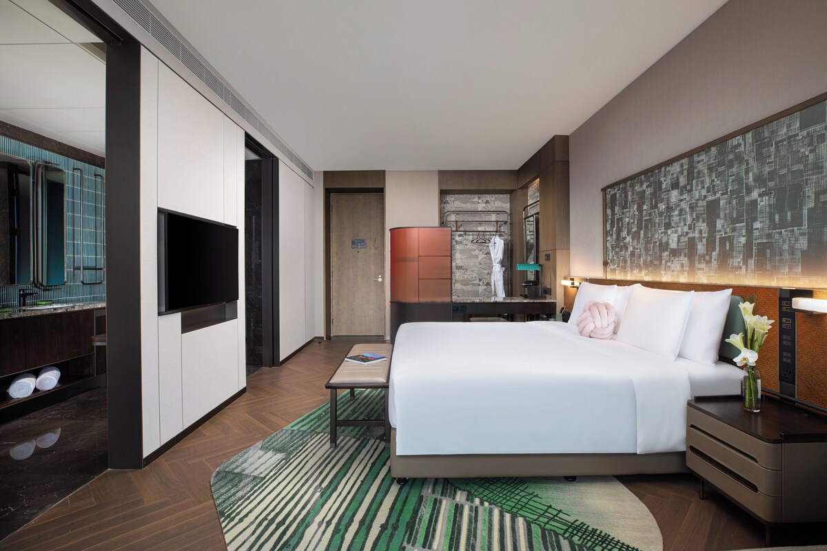 A modern hotel room with a large bed, a flat-screen TV, a desk, and a bathroom with a shower and toilet.