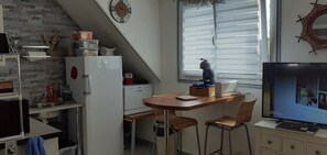 A compact kitchen with a refrigerator, microwave, and sink. There is a small dining table with two chairs. A television is mounted on the wall, and a window with blinds is visible.