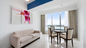 A modern hotel room with a beige sofa, a glass dining table with four chairs, a large window with curtains, and a blue accent wall. - Modern 1 BR Condo near Marlin Azul Restaurant (Marina Mazatlán)