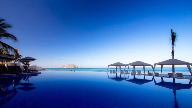 An infinity pool with lounge chairs and umbrellas, overlooking a beach and ocean. - Park Royal Private Junior Suite with Gym & Pool (Marina Mazatlán)