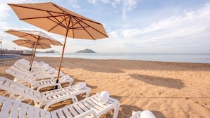 A beach with white lounge chairs and beige umbrellas, a calm sea, and a distant island. - Magnificent Junior Suite with a short Walk to Beach in Mazatlan (Marina Mazatlán)