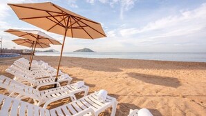 A beach with white lounge chairs and beige umbrellas, a calm sea, and a distant island. - Tasteful Junior Suite with a Terrace and Private Beach Area (Marina Mazatlán)