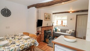 A cozy living room with a fireplace, a dining area with a table set for two, and a kitchen area with a refrigerator. - Barn Cottage (Carnforth)