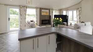 A modern living room with a kitchen island, a flat-screen TV, and a large window with curtains. - Hill View, Lilac Lodge (Wareham)