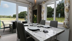 A dining area with a table set for four, large windows with curtains, and a view of a green landscape. - Hill View, Lilac Lodge (Wareham)