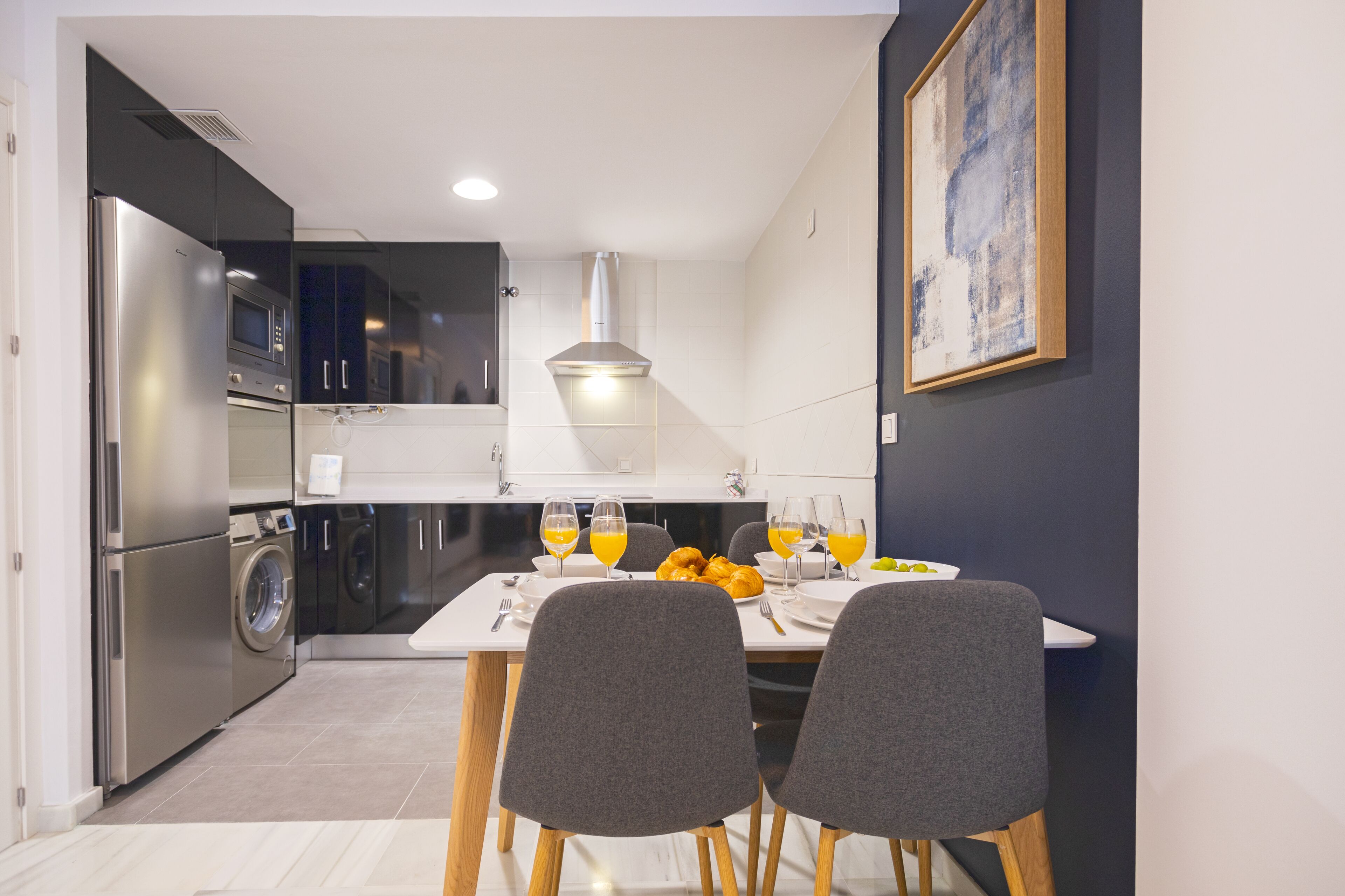 Una cocina moderna con comedor, que incluye un refrigerador, microondas, lavadora y una mesa para dos personas con vasos de jugo de naranja.