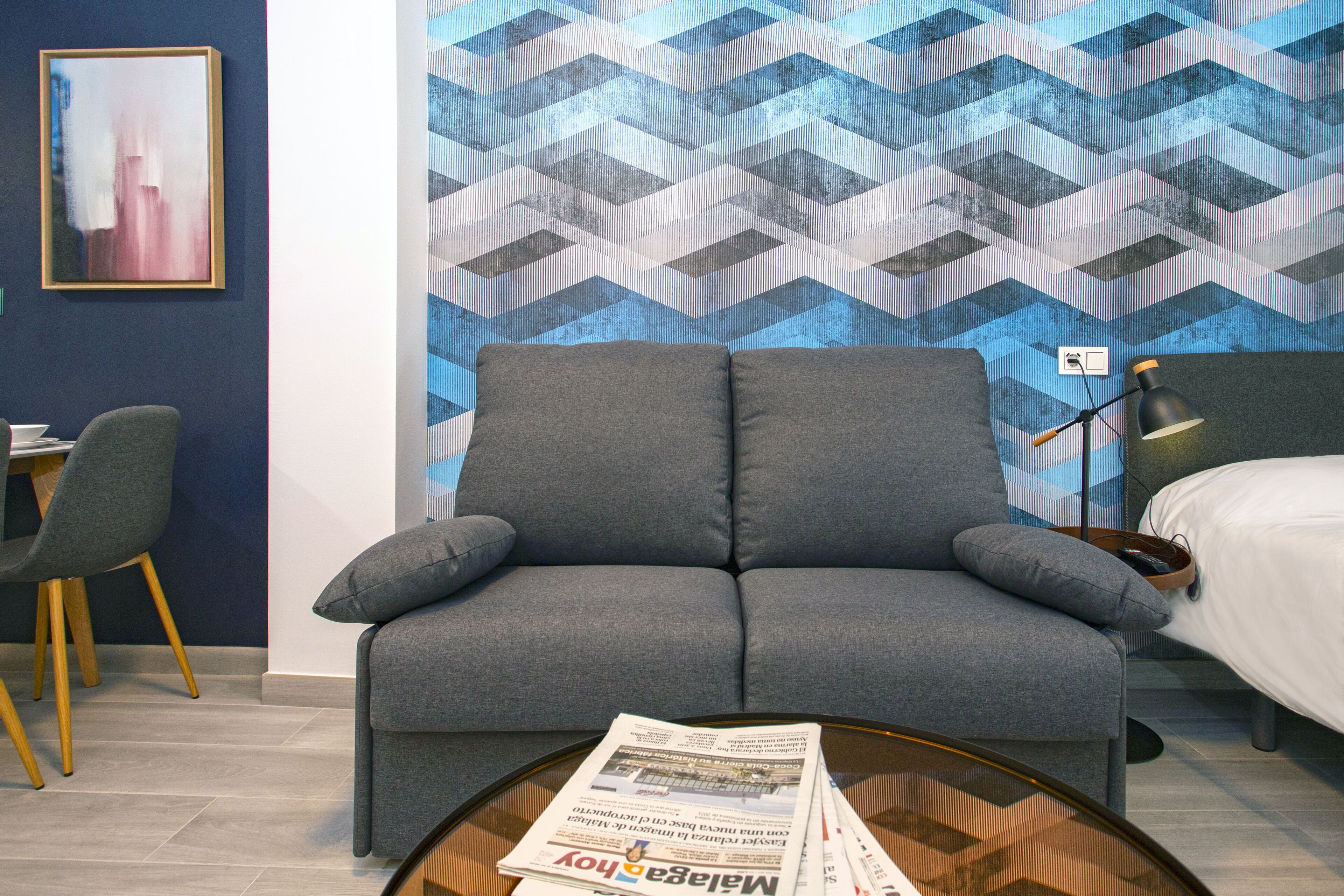 Ein modernes Zimmer mit grauem Sofa, rundem Couchtisch mit einer Zeitung, Schreibtisch und Stuhl sowie einer Wand mit Muster.