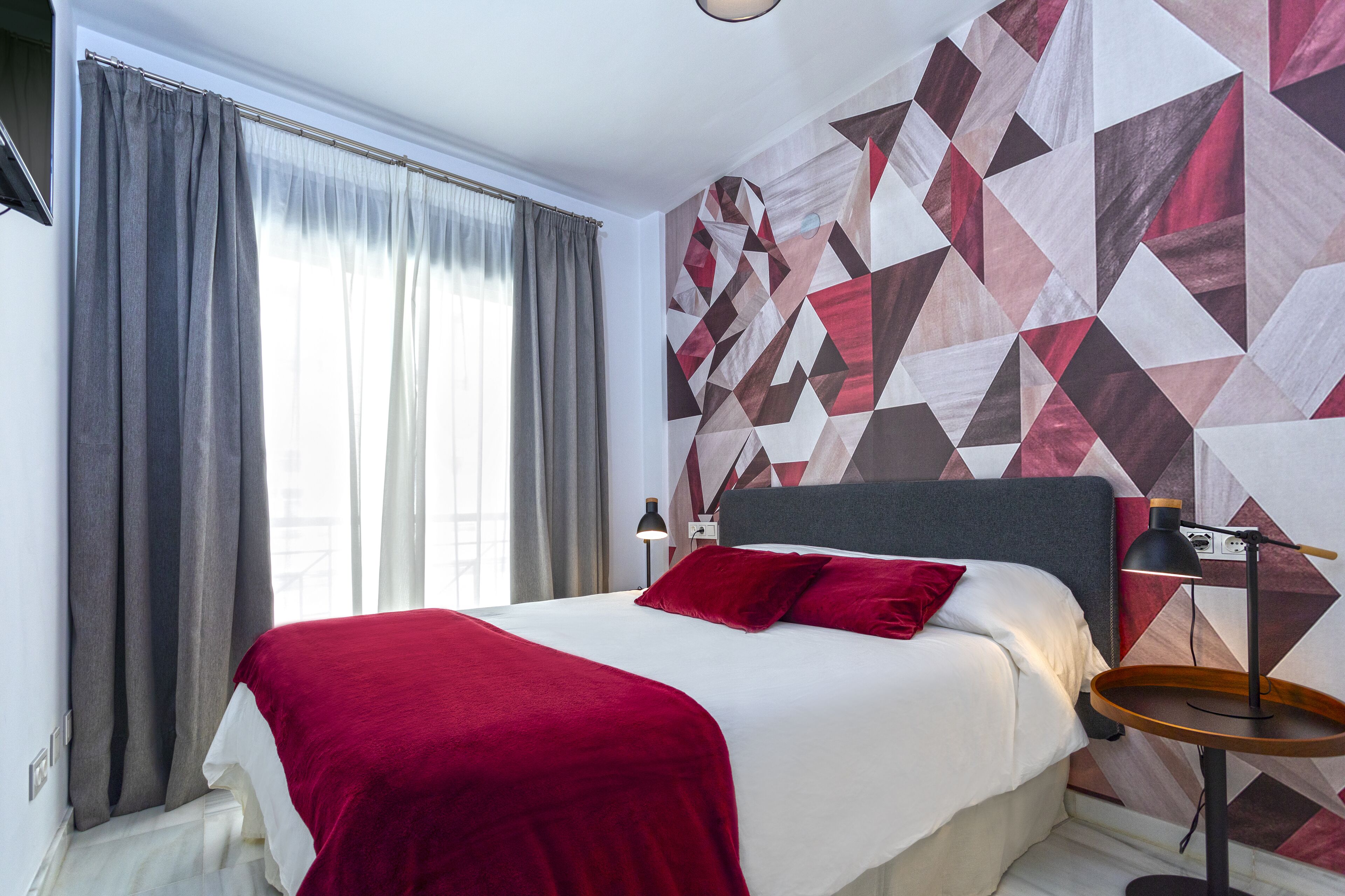 A modern hotel room with a large bed, red bedding, a geometric patterned wall, and a bedside table with a lamp.