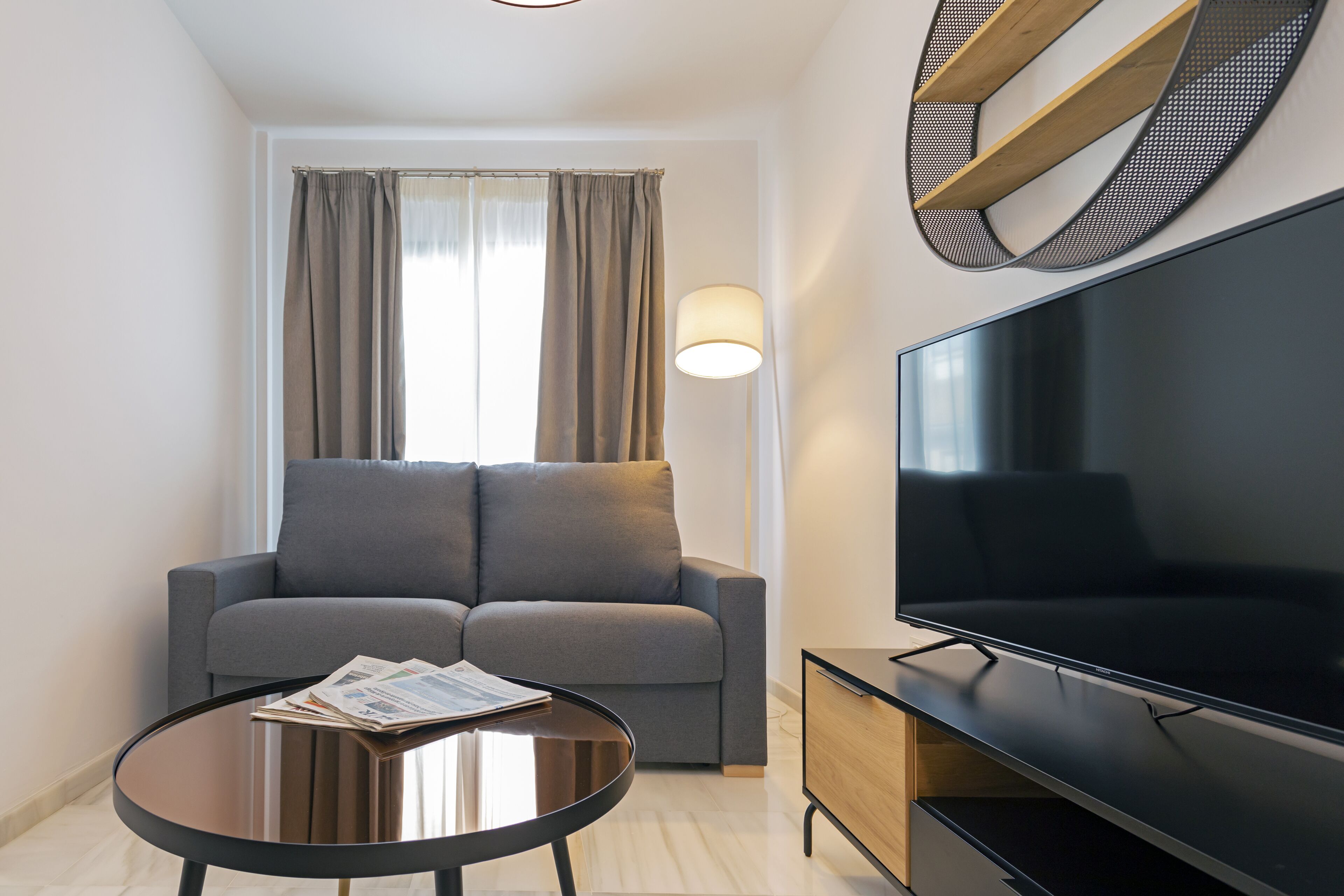 Ein modernes Wohnzimmer mit grauem Sofa, rundem Couchtisch mit Zeitschriften, Flachbildfernseher und einer Wandleuchte.