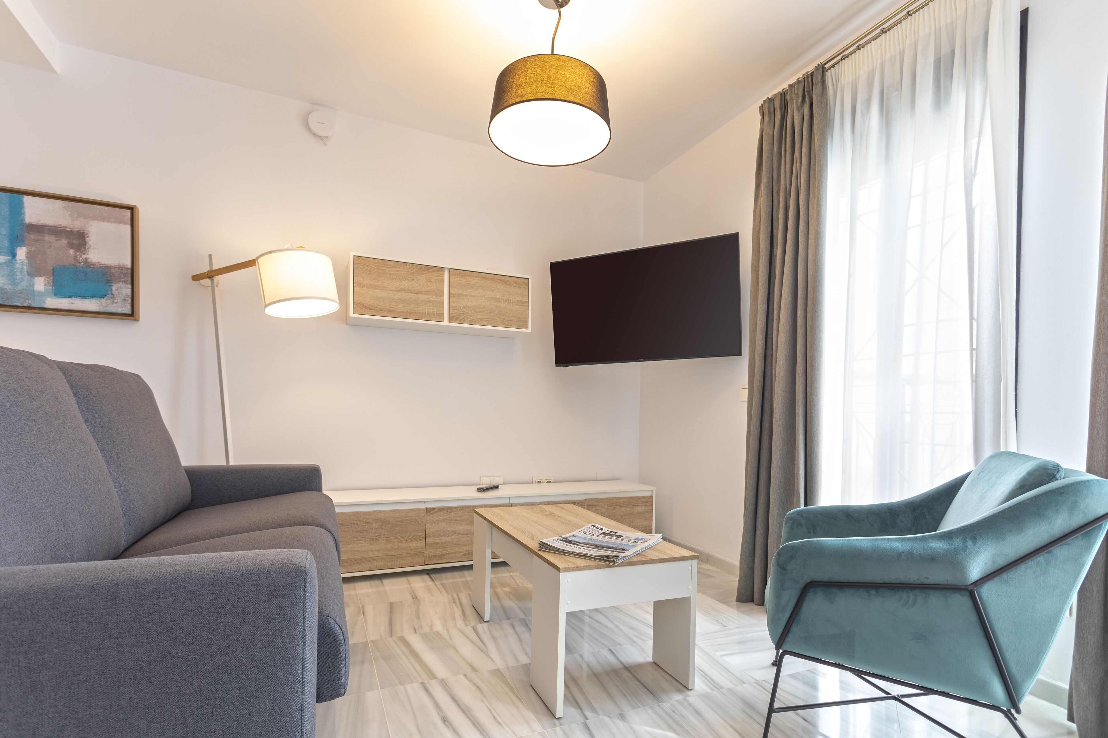 A modern living room with a sofa, a coffee table, a TV mounted on the wall, and a window with curtains.