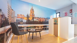 Un hall d’hôtel moderne, agrémenté d’une grande fresque représentant un paysage urbain, d’un comptoir d’accueil et d’un espace détente avec une table et des chaises.