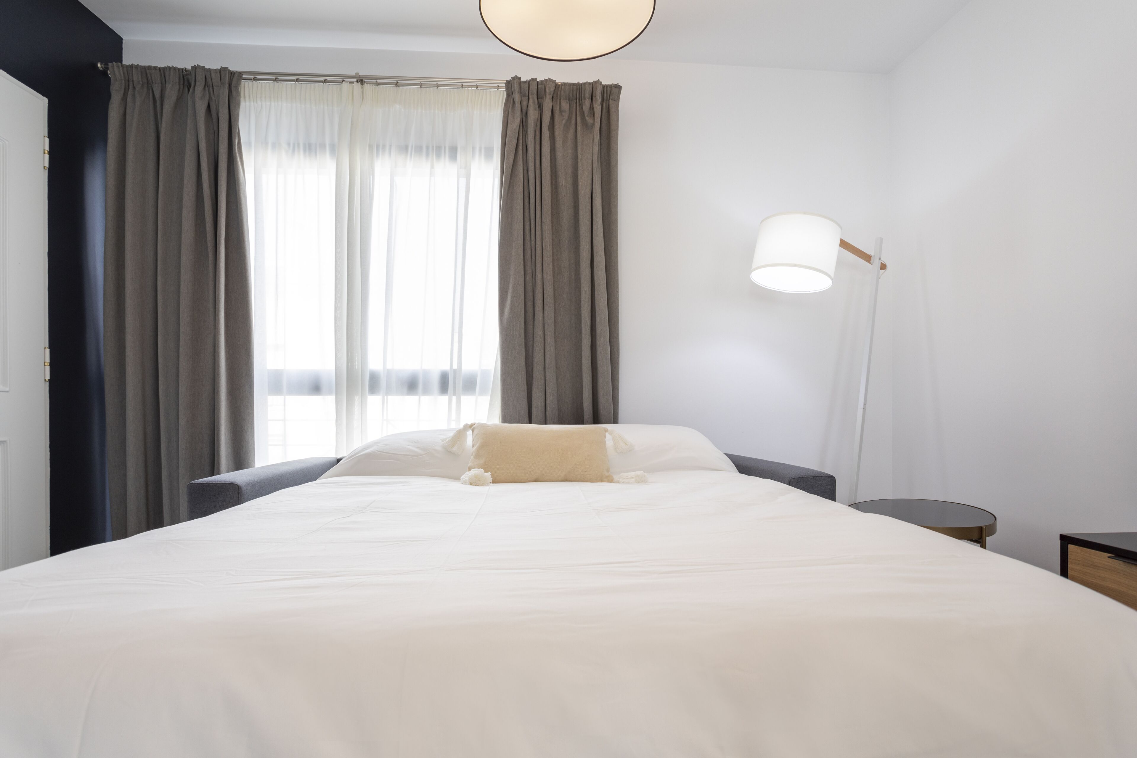 Una cama bien tendida con un cojín color beige, una mesita de noche, una lámpara y una ventana con cortinas.