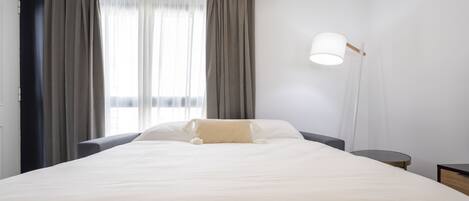 Una cama bien tendida con un cojín color beige, una mesita de noche, una lámpara y una ventana con cortinas.