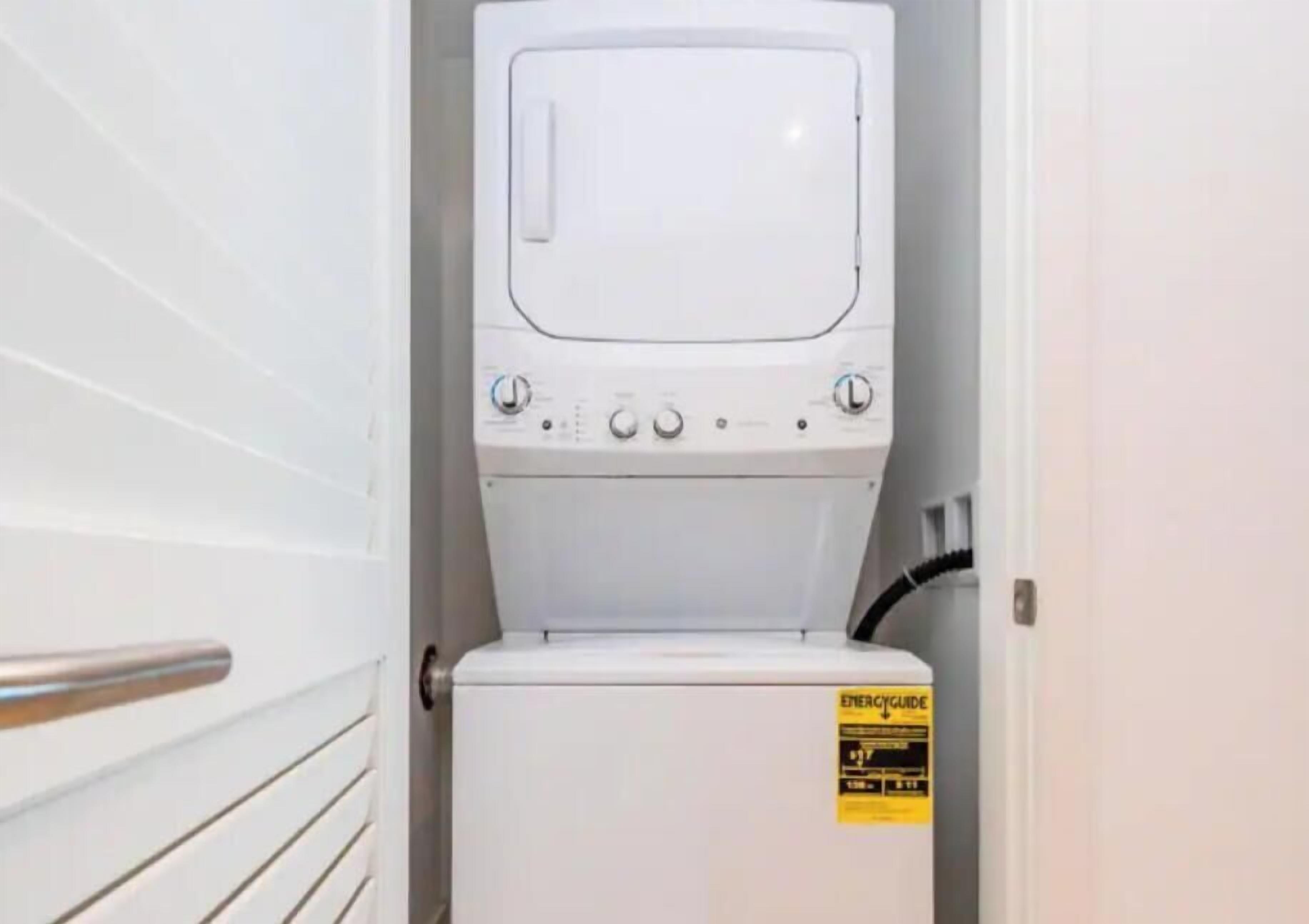 Une petite buanderie blanche, équipée d’un lave-linge et d’un sèche-linge, d’une étiquette d’avertissement de sécurité et d’une poignée métallique sur la porte.