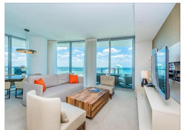 A modern living room with a large sofa, coffee table, and a TV. The room has large windows offering a view of the city and the sea. - Escape to Luxury! Hyde Resort : Ocean Views, Full Amenities & Unbeatable Price! (Hollywood)