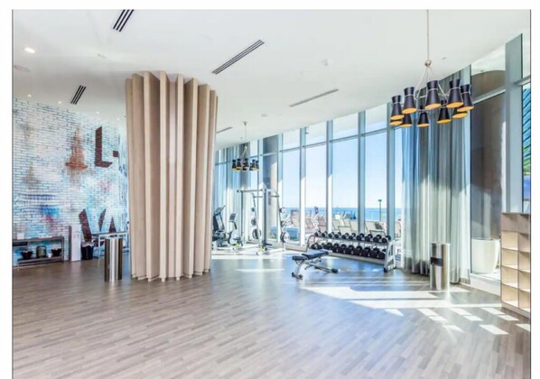 Et moderne fitnesscenter med træningsudstyr, store vinduer og et murmaleri af mursten.