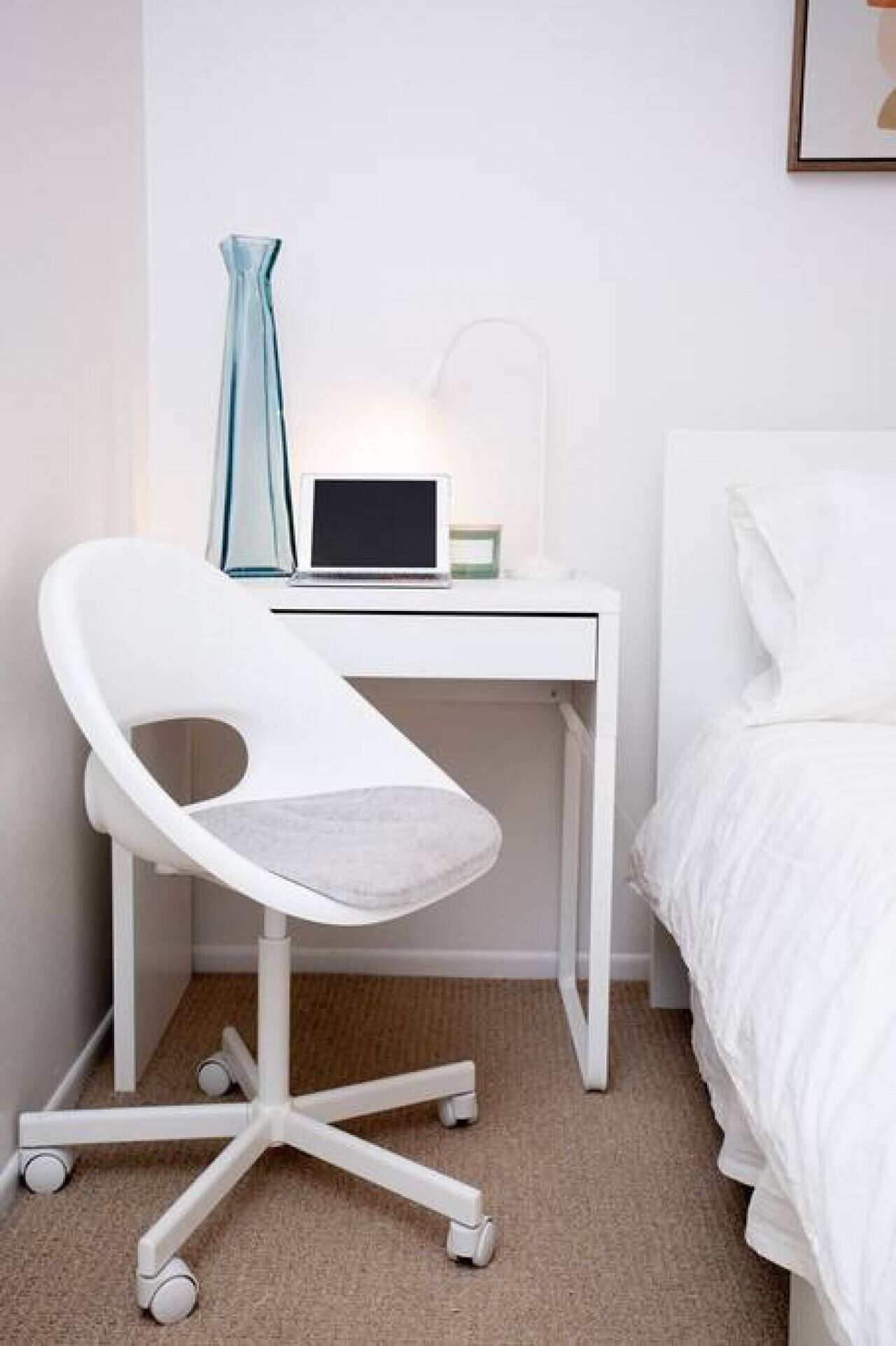 A white desk with a laptop, a modern chair, a bedside table, and a bed with white bedding.