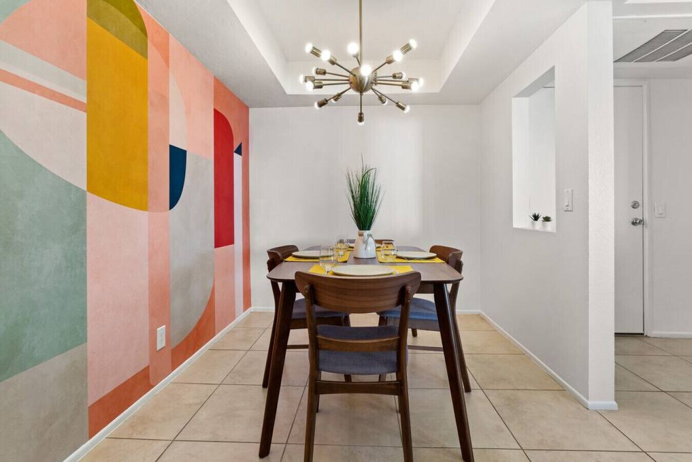 A dining area with a modern chandelier, a table set for four, and a colorful abstract mural on the wall.