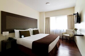 Een hotelkamer met een groot bed, een nachtkastje met een lamp, een stoel en een aan de muur bevestigde televisie.