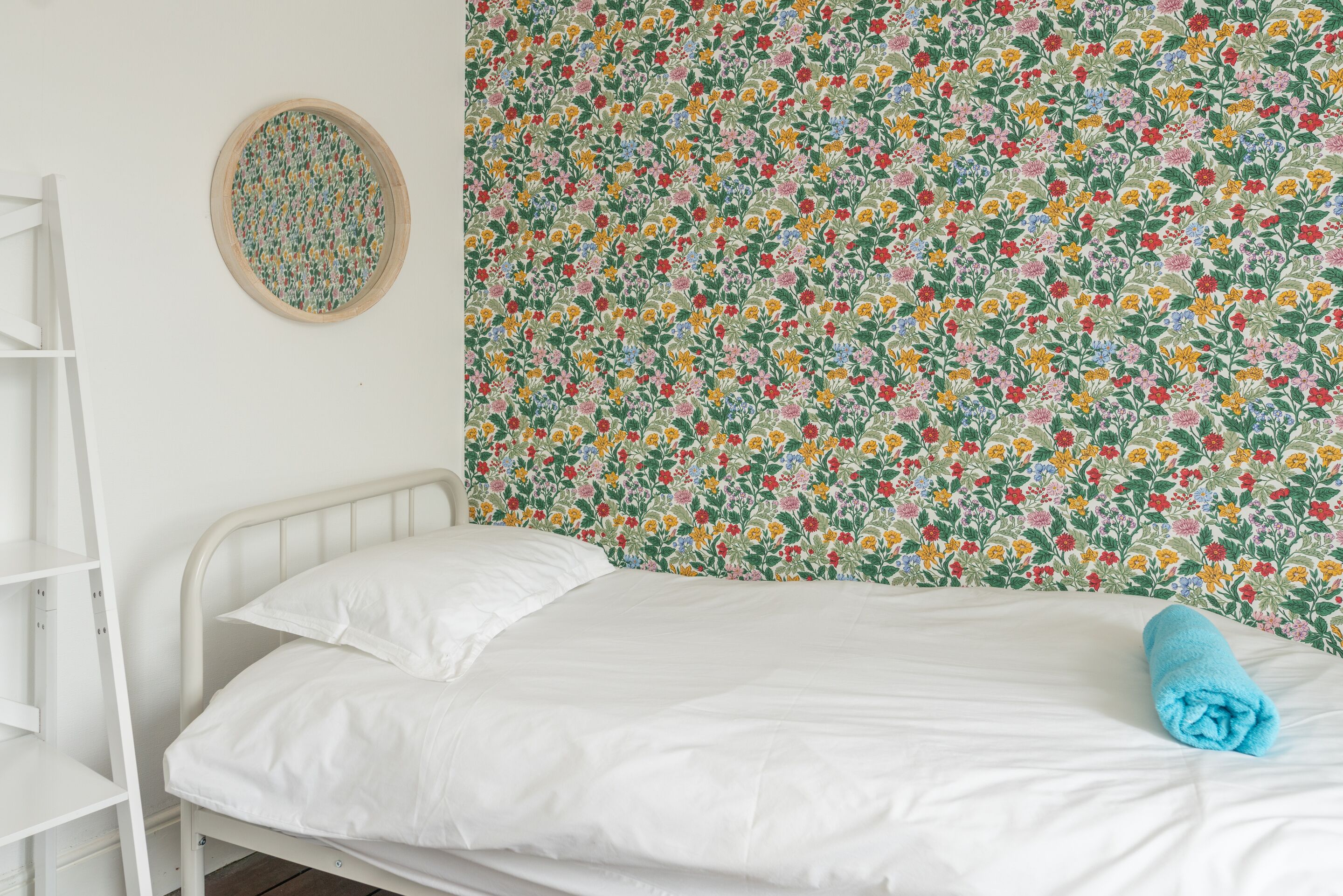 A single bed with white bedding, a blue towel, and a floral-patterned wall.