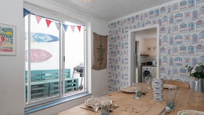 Un coin repas avec une table en bois dressée pour quatre personnes, une vue sur l’extérieur à travers une porte vitrée et une cuisine visible par une porte ouverte.