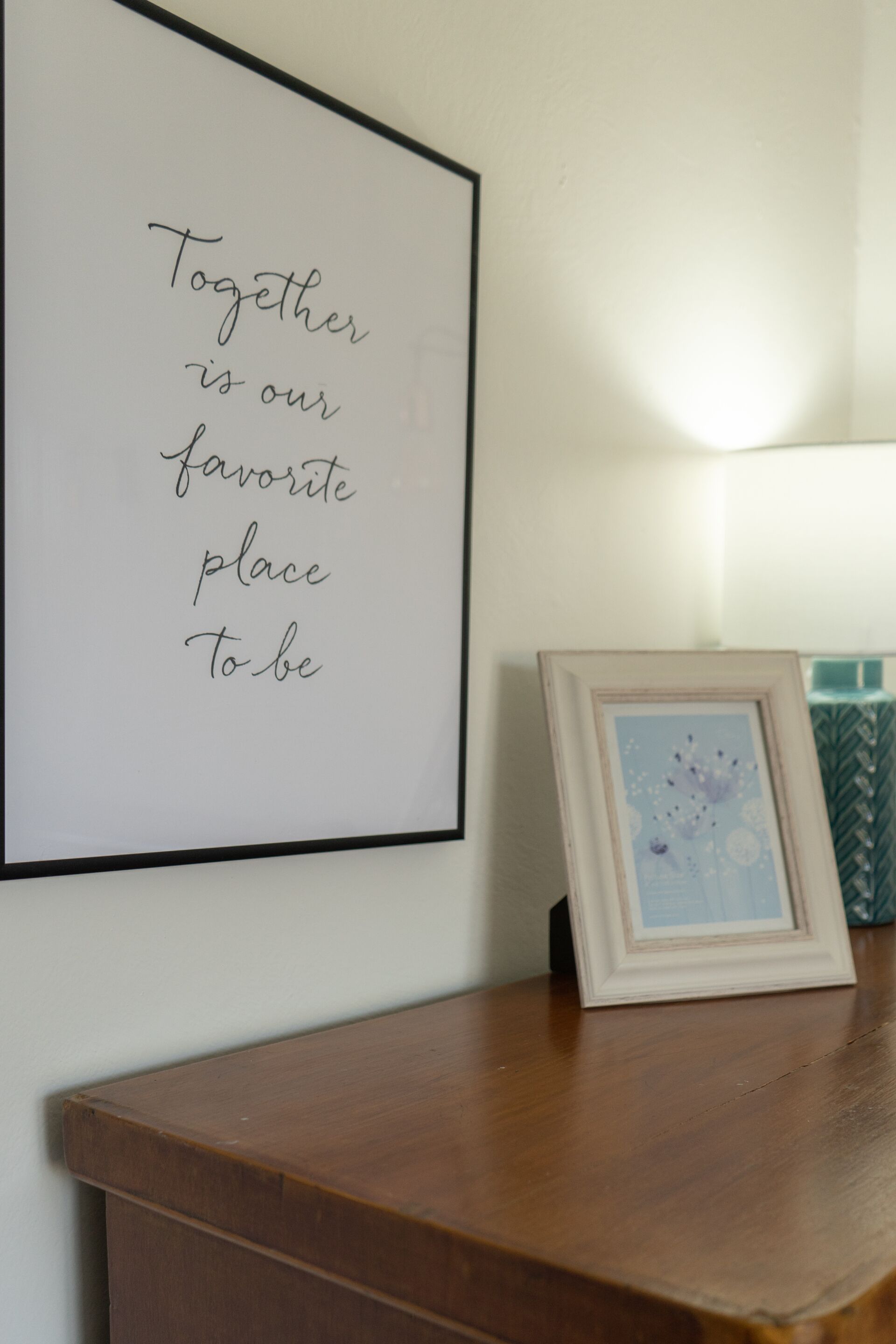 A framed quote on a wall, a framed picture on a wooden table, and a lamp in the background.